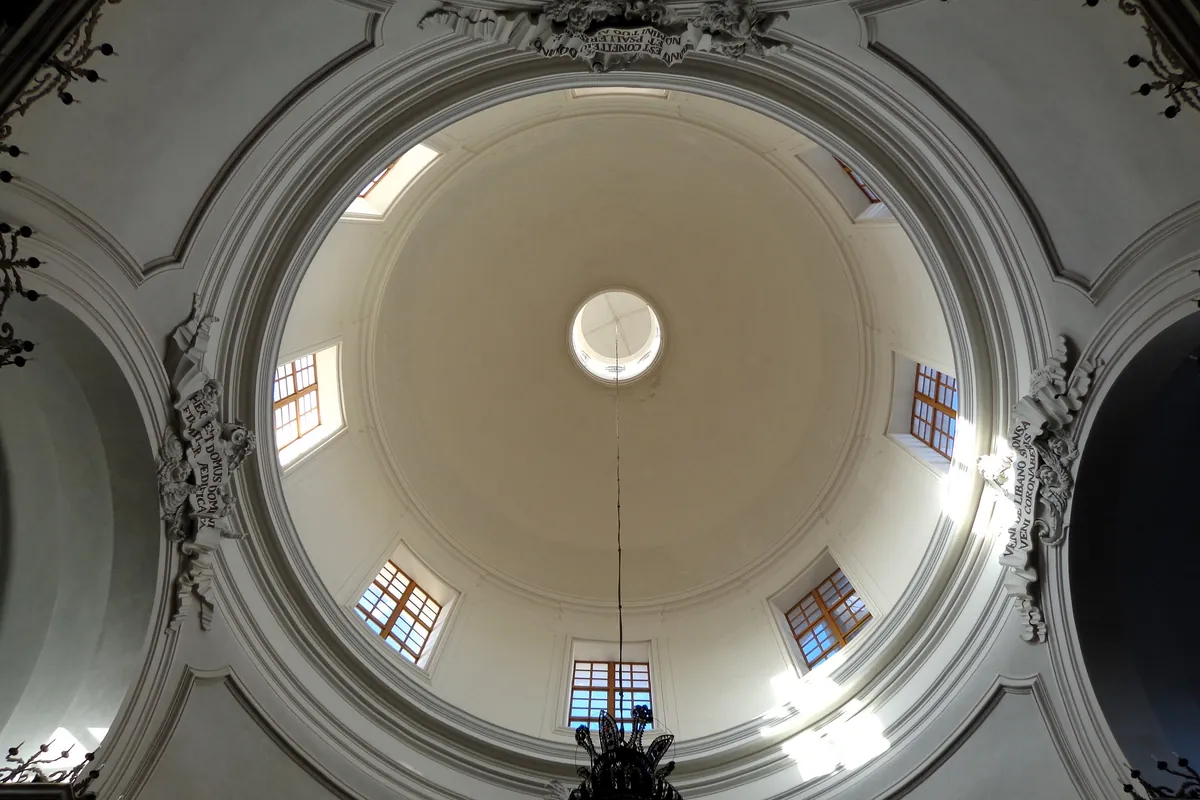 Looking up into the cream dome of the Badia di Sant'Agata, with small windows, white stucco ornament and a chandelier hanging from the centre.