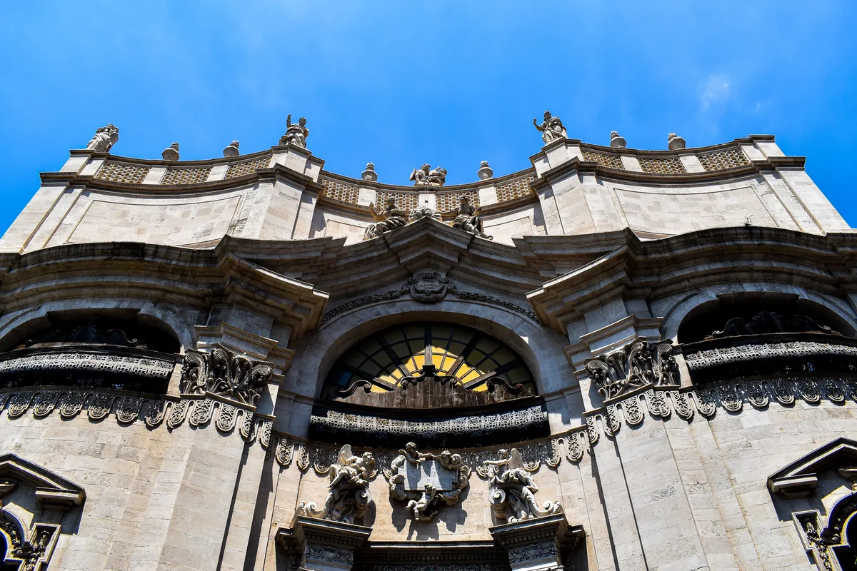 The curving stone facade of the Badia di Sant'Agata seen from below, with carved cornices, statues and a large window above the door against a blue sky.