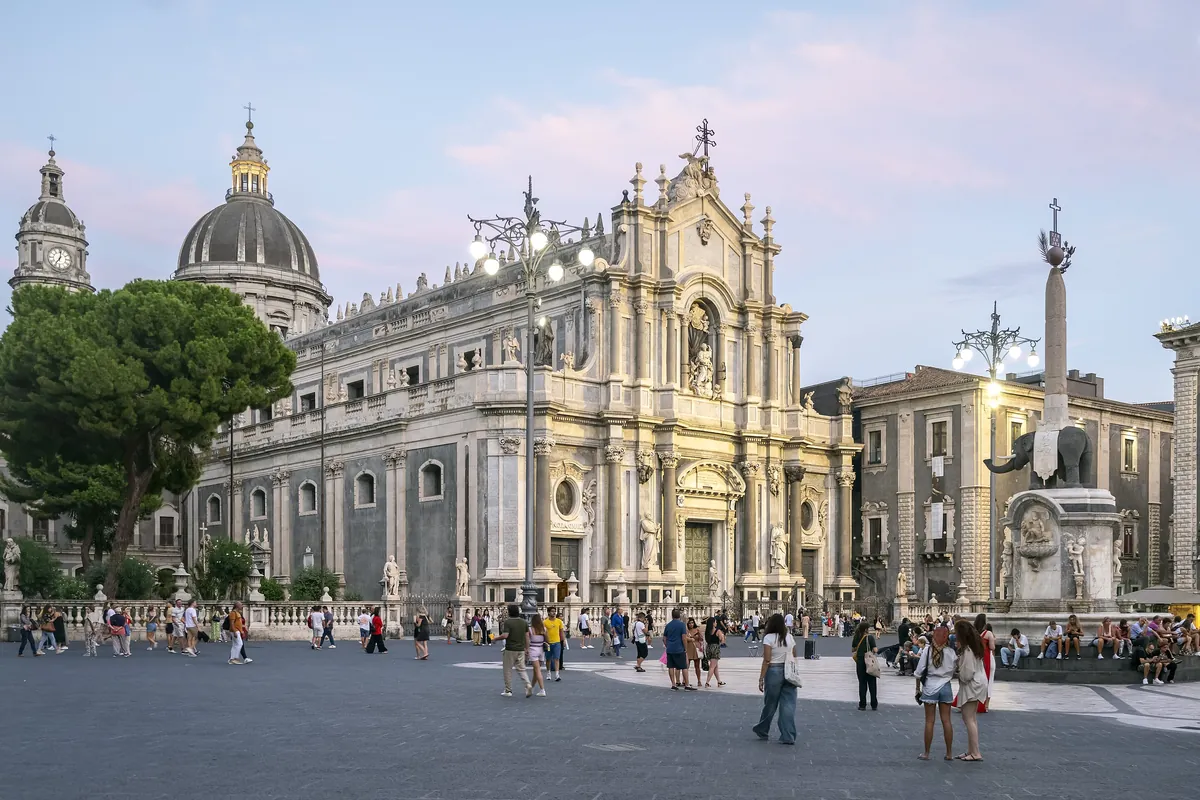 Piazza del Duomo in Catania at dusk, with the cathedral facade, the lava-stone elephant fountain and its obelisk, and people crossing the square.